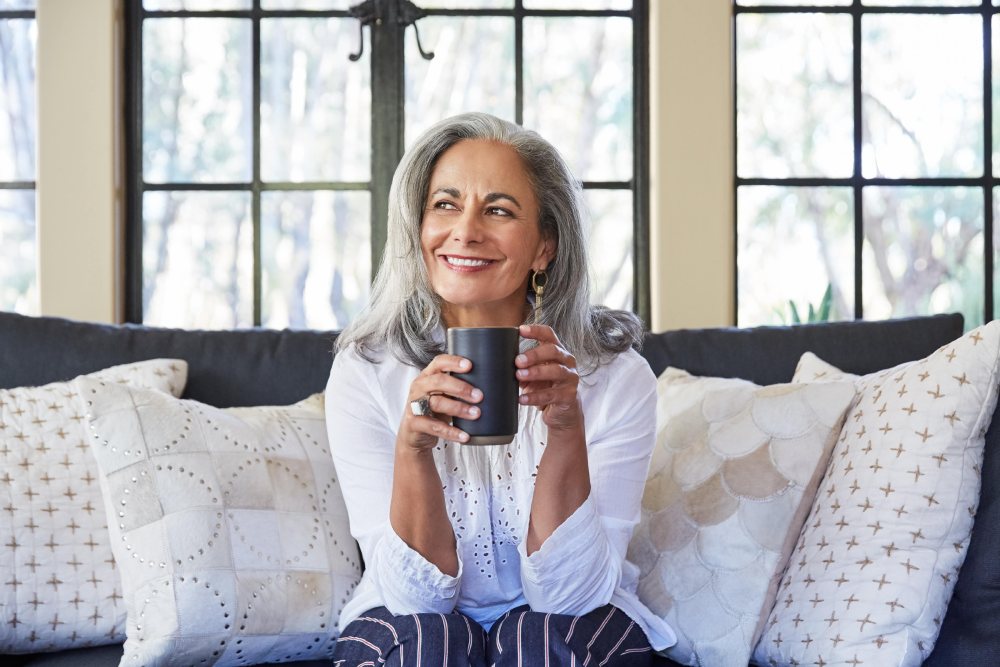 Una mujer mayor sentada en un sofá sosteniendo una taza de café y sonriendo.