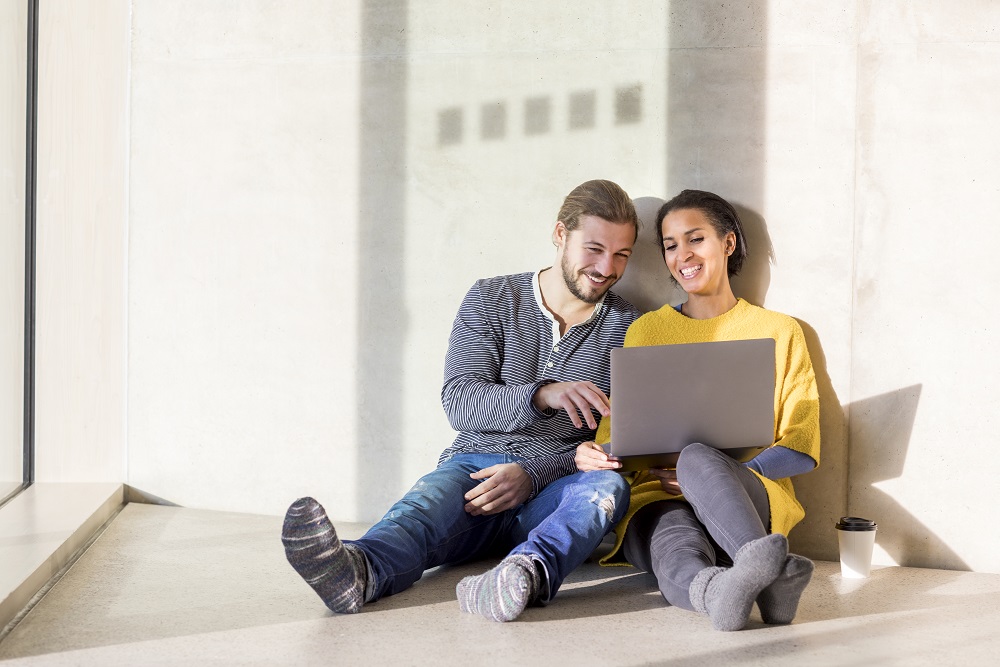 On-page, 2024. Laughing young couple sitting on the floor using a laptop