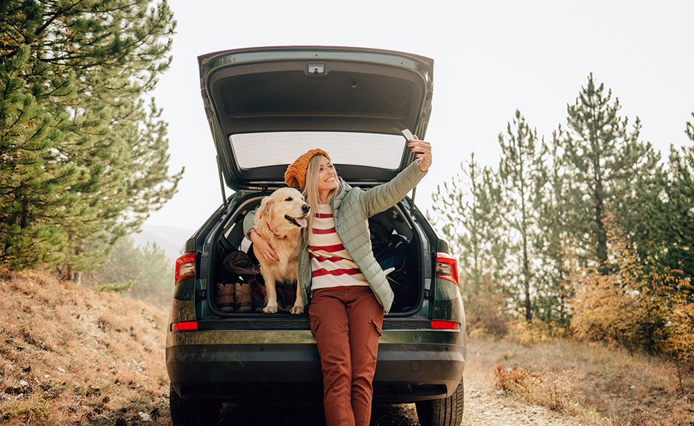 Woman sitting next to her dog in an open van trunk taking a selfie.