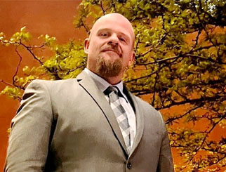 A man in a tan suit standing in front of a tree and building.